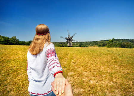 Beautiful Ukrainian girl in white ethnic shirt leading man by hand to wooden wind mill at architecture museum in Pirogovo. Kiev, Ukraineのeditorial素材