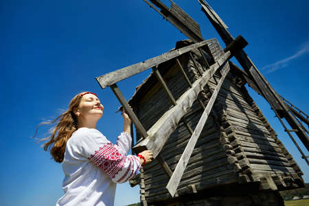 Beautiful Ukrainian girl near wooden wind mill in white ethnic shirt at national architecture museum in Pirogovo. Kiev, Ukraineのeditorial素材