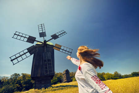Beautiful Ukrainian girl dancing near wooden wind mill in white ethnic shirt at national architecture museum in Pirogovo. Kiev, Ukraineのeditorial素材