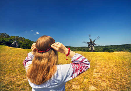 Beautiful Ukrainian girl wearing flower wreath at wooden wind mills background in white ethnic shirt at national architecture museum in Pirogovo. Kiev, Ukraineのeditorial素材