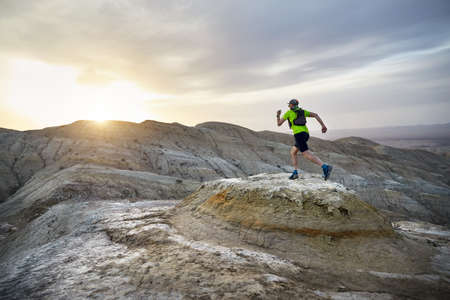 Runner athlete with backpack running on the wild mountains in the desert at sunsetの写真素材