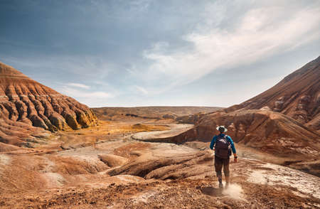 Tourist with backpack walking at the dusty trail on surreal red mountains against blue sky in desert park Altyn Emel in Kazakhstanの写真素材