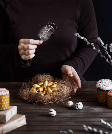 Woman holding golden chocolate eggs in the nest near cakes and presents on Easter holidays