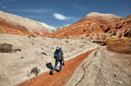 Tourist with backpack and camera walking at the dusty canyon on surreal red mountains against blue sky in the desertの写真素材