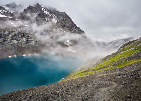 Beautiful landscape of turquoise Ala-Kul Lake in the Tien Shan mountains with white foggy clouds and small tourist in red in Karakol national park, Kyrgyzstanの写真素材