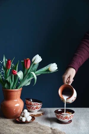 Kazakh woman pouring milk in Kazakh tea bowl kese with ornament near kurt and tulips on the table during Nauryz festival at dark blue background.の写真素材