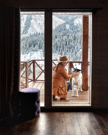 Woman in hat is playing with her dog at wooden house in the mountain resort at winterの写真素材