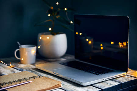 Cozy workspace with laptop, note with pencil and cup of coffee, plants and yellow glowing garlandat stone grey wall background. Working from home concept.の写真素材