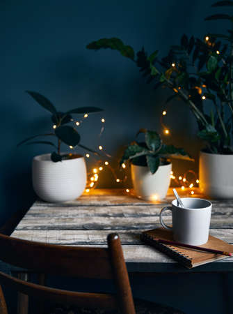 Cozy workspace with note book with and cup of coffee at wooden table with plants and yellow glowing garland at stone grey wall background. Working from home concept.の写真素材