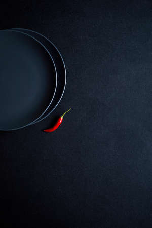 Minimalist composition of red chili pepper near two ceramic plates at black tableの写真素材