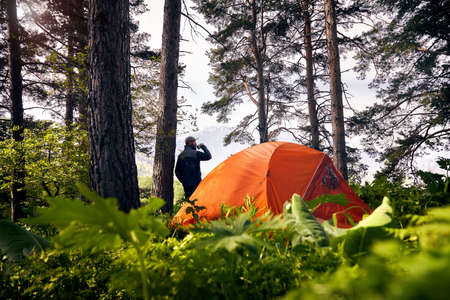 Bearded tourist is drinking coffee near orange tent in the lush forest at mountains  
