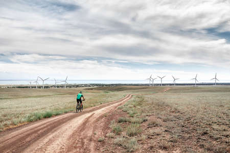 Man on mountain bike rides on the road to the wind turbines farm near the lake Against blue cloudy sky. Ecological and zero waste lifestyle concepts.の写真素材
