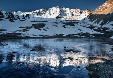 Beautiful scenery of the mountain lake with ice and reflection of snowy mountains at sunriseの写真素材