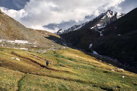 Small tourist with backpack is walking in the green mountain valley with snowy peaks and cloudy sky backgroundの写真素材