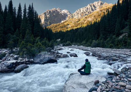Young man doing yoga meditation in Padmasana sitting position on the rock near the river with...