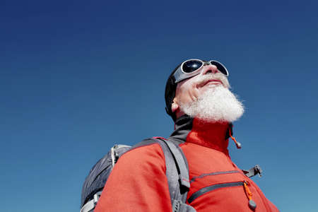 Portrait of climber with long white beard in red jacket against blue clear sky in the mountainsの写真素材