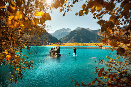 Man floating on a SUP board at mountain lake near yellow forest in autumn time. Adventure at Stand up paddle boarding.の写真素材