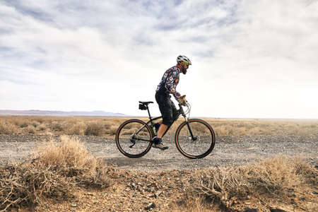Rider with beard rides his mountain bike in the desert road at sunny weather in Kazakhstan. Extreme...