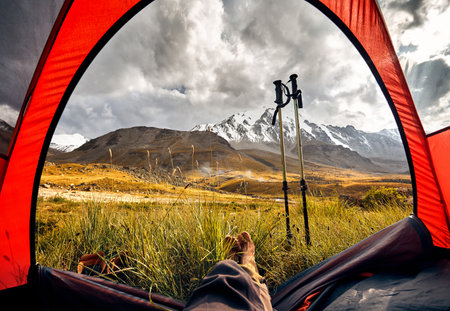 View from inside a tent and hikers legs to the mountains with glacier and trekking poles in...