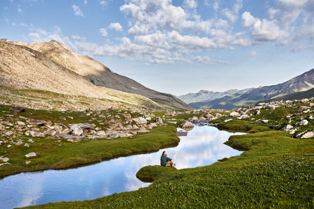 Man sitting and resting near clear mountain river with reflection blue cloudy sky