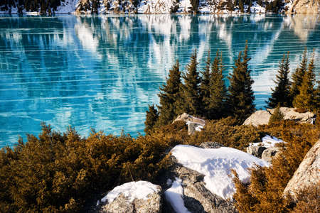 Beautiful landscape of mountain lake with blue ice and spruce forest at winter timeの写真素材