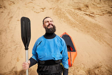 Portrait of Old man with black beard and paddle in blue jacket smiling near orange raft sport boat at sand dune backgroundの写真素材