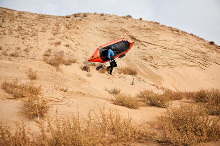 Old man with black beard in blue jacket holding orange raft sport boat at sand dune in the desert background. Adventure and travel concept.の写真素材