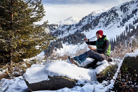 Man with beard in red hat pouring hot tea from cup in the spruce forest against winter mountain background.の写真素材