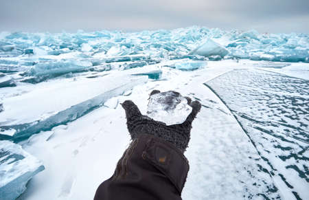 Open hand in glove holding small transparent piece of Ice at frozen lake with snowの写真素材