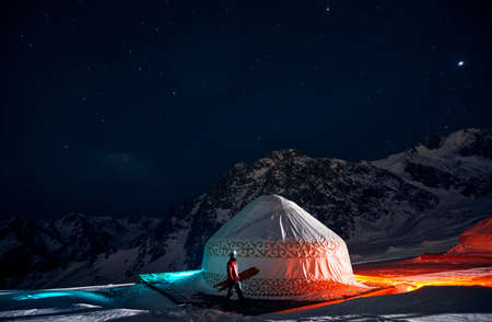 Man walking with split snow board near Yurt nomadic house at Ski resort Shymbulak in Almaty, Kazakhstan. Winter night astrophotography with stars and color lights backcountry rider.の写真素材