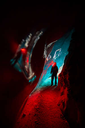 Silhouette of Alpinist in helmet with red glow headlamp in deep narrow glacial ice cave with cyan...