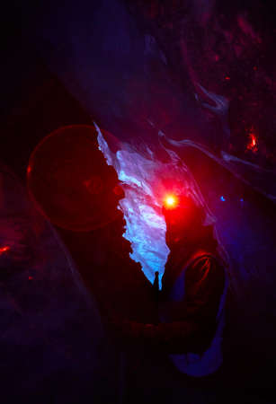 Bearded Alpinist in helmet with red glow headlamp in deep narrow glacial ice cave with blue lights...