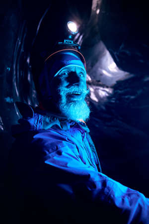 Portrait of Old Alpinist with grey beard in helmet with glow headlamp smiling in deep dark glacial ice cave with blue lights explore winter mountain glacier in Kazakhstanの写真素材