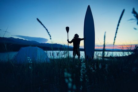 Man in silhouette with paddle and SUP board near tent camping near lake at sunset in Kazakhstan. Stand up paddle boarding outdoor active recreation in nature.の写真素材