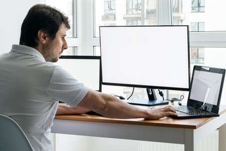 A man in a white t-shirt is sitting at a workplace with two laptops and a monitor near the window. Remote work from home in quarantineの写真素材