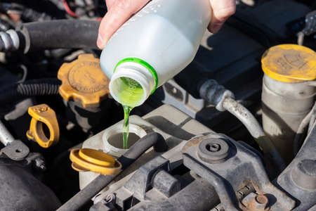 Hand with bottle pouring antifreeze coolant into the expansion tank. Dusty details of a flat-four (boxer) car engine compartment under the open hood. Closeup view on a sunny dayの写真素材