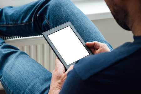 A man in a blue t-shirt and jeans holds an e-reader electronic book with white blank mockup screen in his hands. Reading books at home by the window. Selective focusの写真素材