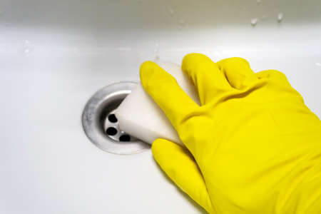 A hand in a yellow rubber glove with a melamine sponge rubs the wet white bath or the sink near the drain hole. Cleaning in a modern bathroom. Selective focus. Closeup viewの写真素材