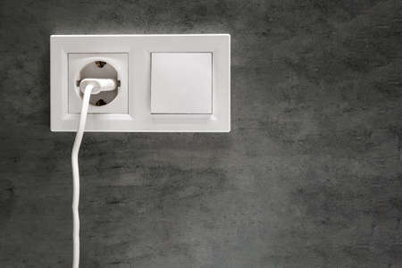 A closeup view of a group of white european electrical outlet with a plug inserted into it and a switch located on a concrete gray wall in a modern bathroom with copy spaceの写真素材