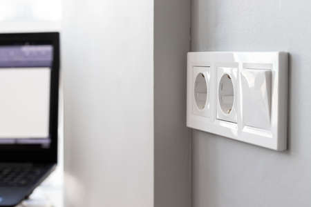 Group of white european electrical outlets and a switch located on a gray wall in a bright modern room with a blurred working laptop by the window. Selective focus. Closeup viewの写真素材