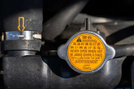 Orange radiator cap with warning labels in different languages: never open when hot, and blurred car radiator hose with yellow dotted arrow under the open hood of a car. Closeup viewの写真素材