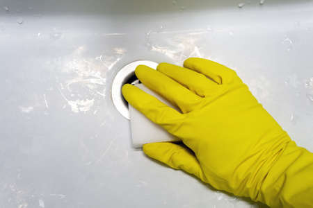 A hand in a yellow rubber glove with a melamine sponge rubs the wet dirty white bath or the sink near the drain hole. Cleaning in a modern bathroom. Selective focus. Closeup viewの写真素材