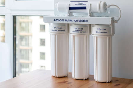 Four stages water filtration system for the home with: coconut postcarbon filter, granular activated and block carbon on a wooden table against a blurred window background. Reverse osmosis systemの写真素材