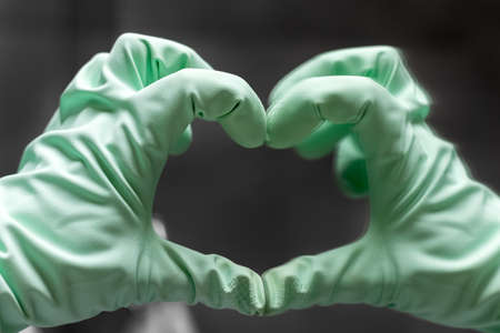 A hand in a mint green rubber glove shows a heart sign with its reflection in the mirror against a gray blurred background. Selective focus. Closeup viewの写真素材