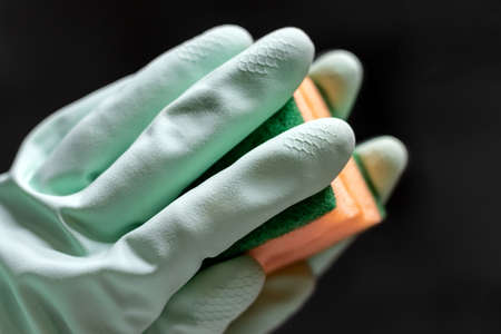 A hand in a mint green rubber glove wipes a mirror in a dark room with an orange sponge. Closeup view. Selective focusの写真素材