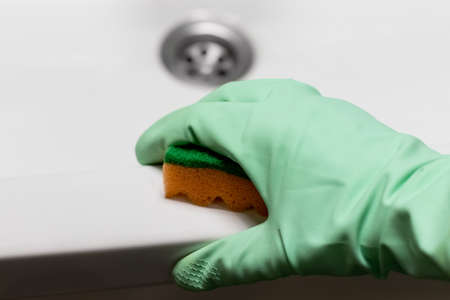 A hand in a mint green rubber glove wipes the surface of a white bath or sink with an orange sponge. Cleaning in a modern bathroom. Selective focus. Closeup viewの写真素材