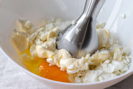 Cottage cheese, butter, egg and sugar in a white bowl - ingredients for mixing with a hand blender. The process of making curd bagels or croissantsの写真素材
