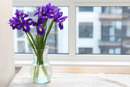 Bouquet of purple irises in a clear glass vase on a linen tablecloth on a wooden table by the window in a modern bright kitchen against a blurred backgroundの写真素材