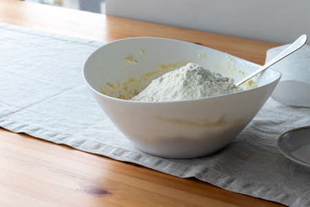 A white bowl with cottage cheese dough, on which a hill of flour is poured, stands on a linen tablecloth on a wooden table near the window. The process of making curd bagels or croissantsの写真素材