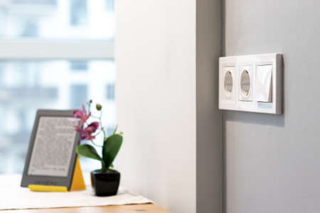 Group of white european electrical outlets and a switch located on a gray wall in a light modern kitchen with flower and e-book on the table by the window. Selective focusの写真素材